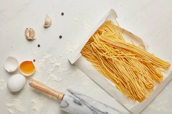 Flat lay with raw chicken eggs, uncooked pasta, flour and rolling pin on white marble tabletop — Stock Photo