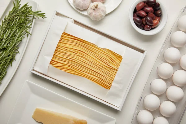 Vista superior de la disposición de macarrones sin cocer, ajo, romero y queso para cocinar pasta en la mesa blanca - foto de stock