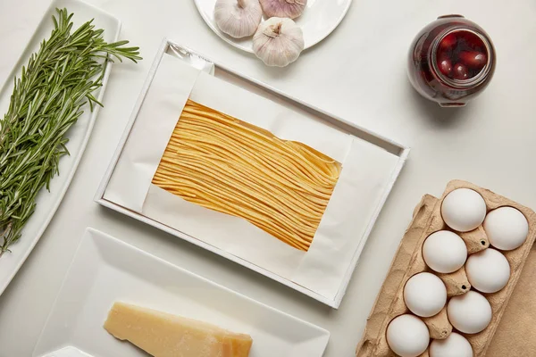 Vista superior de la disposición de macarrones sin cocer, ajo, romero y queso para cocinar pasta en la mesa blanca - foto de stock