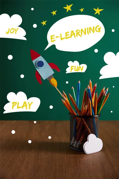 Colorful pencils, rocket, cloud sign on wooden table with chalkboard on background with play, joy, fun and e-learning words — Stock Photo