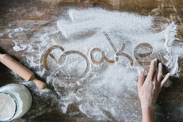 Обрезанный снимок женщины, пишущей слово BAKE с пальцем на деревенском деревянном столе, покрытом мукой — стоковое фото