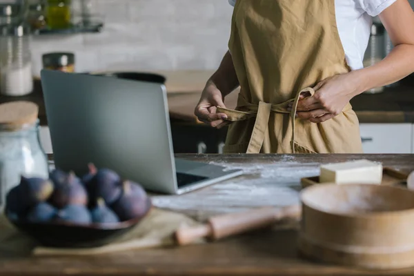 Tiro recortado de la mujer con el ordenador portátil y pastel ingredientes en rústico mesa de madera atar delantal - foto de stock