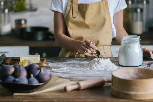 Tiro cortado de mulher em avental em pé na frente da mesa de madeira rústica com ingredientes de torta e pilha de farinha — Fotografia de Stock