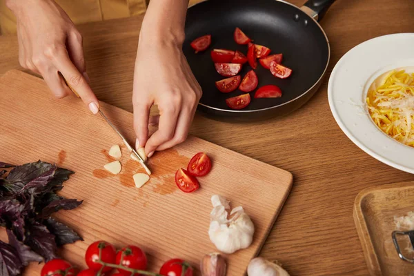 Tiro recortado de mujer cortando ajo y tomates para pasta en la mesa de la cocina - foto de stock