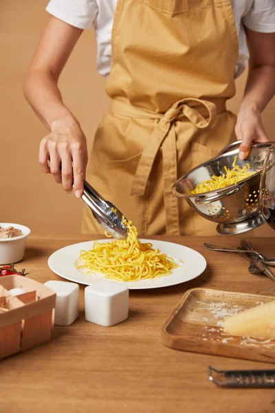 Cropped shot of woman in apron putting spaghetti on plate with tongs from colander — Stock Photo