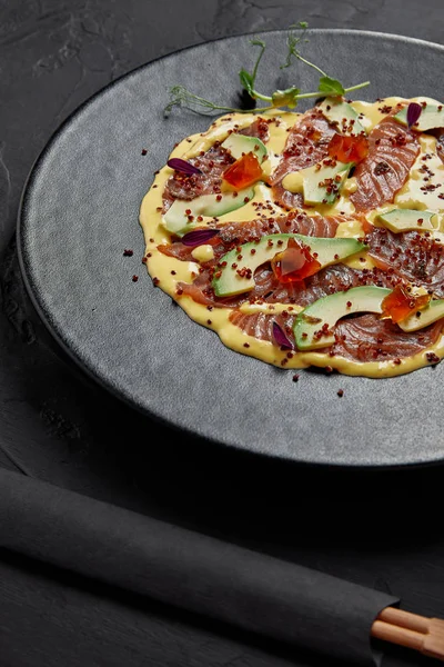 Nahaufnahme von köstlichen Fischscheiben und Avocado in Sauce auf schwarzem Teller — Stockfoto