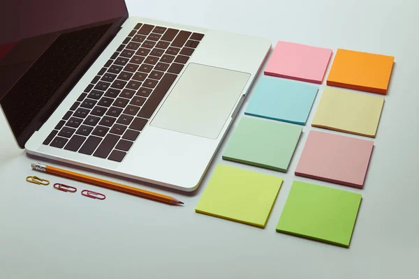 Portátil, conjunto de pegatinas de papel de colores, lápiz y clips de papel en la mesa blanca - foto de stock