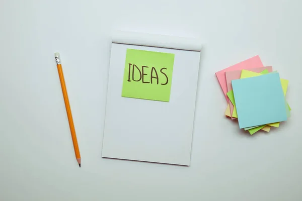Elevated view of paper sticker with word ideas in notebook, pencil and pile of note papers on white tabletop — Stock Photo