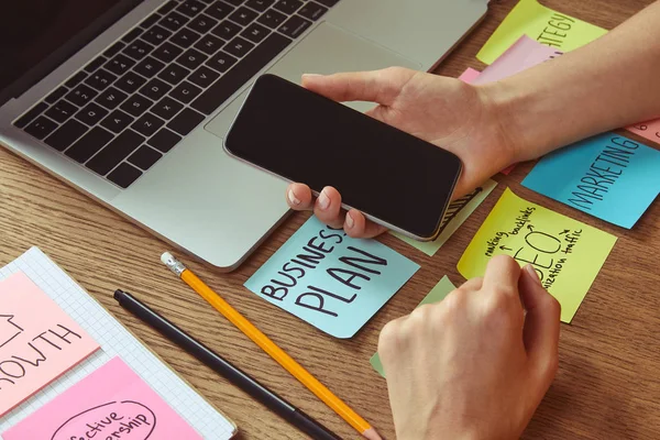 Cropped image of woman holding smartphone with blank screen at table with business plan sticker — Stock Photo