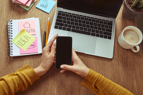 Cropped image of woman holding smartphone with blank screen at table with laptop and coffee — Stock Photo