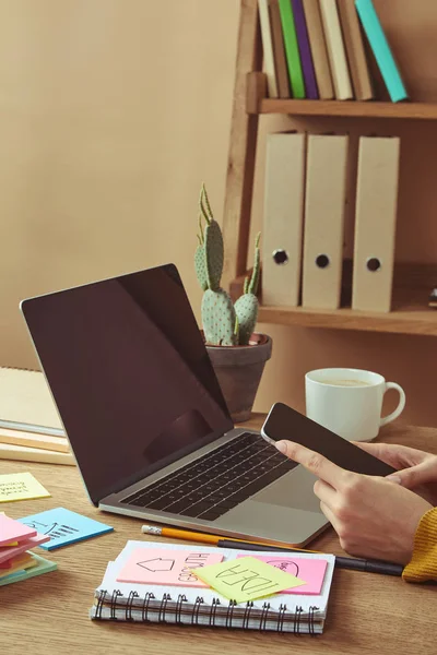 Imagem cortada de mulher segurando smartphone com tela em branco à mesa com laptop e adesivos — Fotografia de Stock