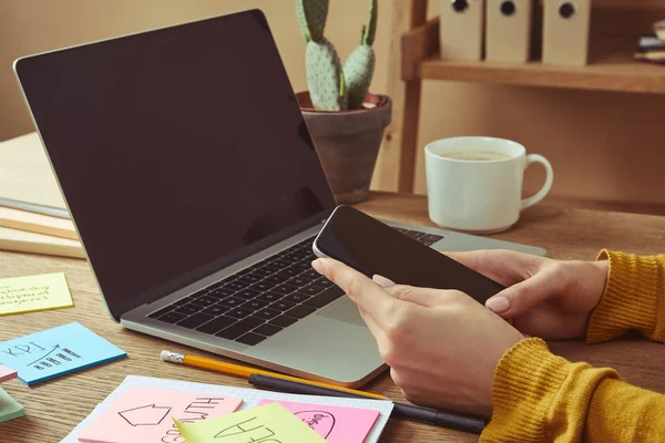 Cropped image of girl holding smartphone with blank screen at table with laptop and stickers — Stock Photo