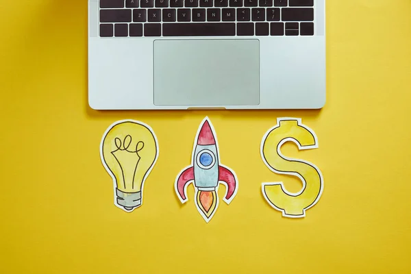 Top view of laptop, paper lightbulb, paper rocket and dollar sign on yellow surface — Stock Photo