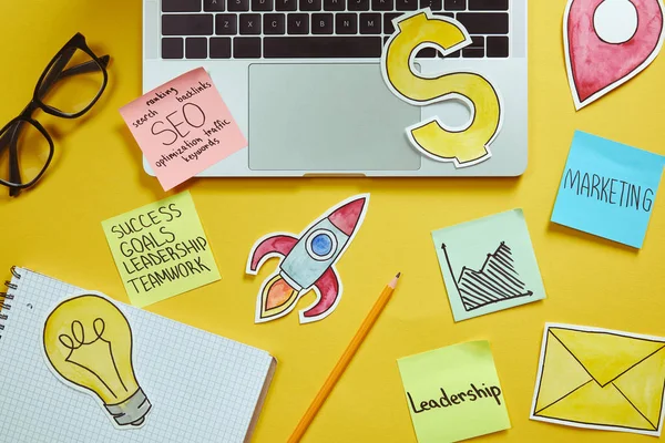 Вид на ноутбук, бумажные вывески и бумажные наклейки с бизнес-планом на желтой поверхности — стоковое фото