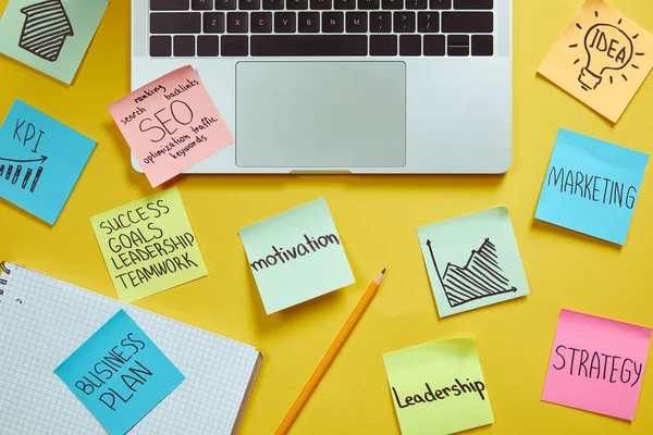 Top view of laptop and paper stickers with business plan on yellow surface — Stock Photo