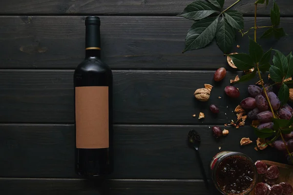 Верхний вид бутылки вина с пустой этикеткой, виноград, орехи и джем на деревянной поверхности — стоковое фото