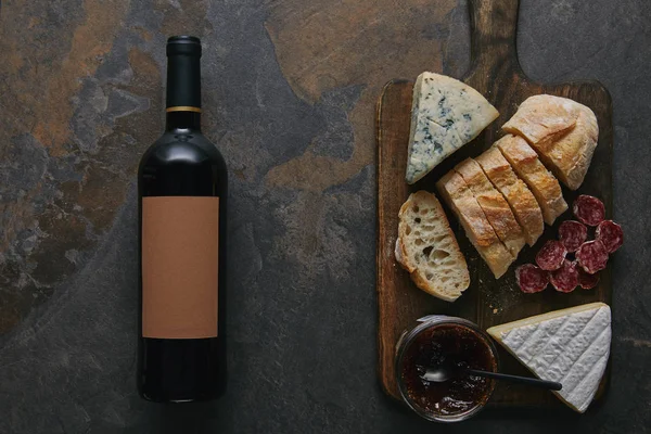 Vue du dessus de la bouteille de vin avec étiquette vierge, pain tranché, salami et fromage sur planche à découper en bois — Photo de stock