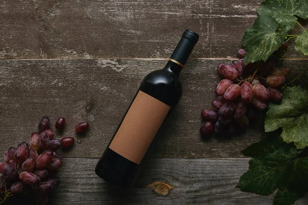 Vista superior de garrafa de vinho com rótulo em branco e uvas maduras frescas com folhas verdes na mesa de madeira — Fotografia de Stock