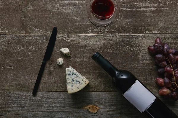 Vista superior de la botella de vino con etiqueta en blanco, vidrio con vino tinto, queso, uvas y cuchillo en la mesa de madera — Stock Photo