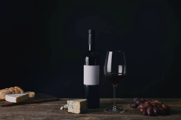 Bouteille de vin avec étiquette vierge, verre de vin rouge, fromage et raisins sur table en bois — Photo de stock