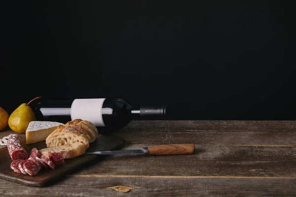 Botella de vino con etiqueta en blanco, peras, deliciosos aperitivos y cuchillo en la mesa de madera - foto de stock