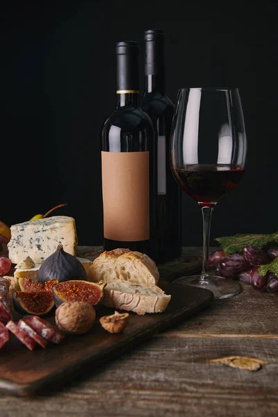 Vista de perto de garrafas e copo de vinho tinto, frutas e delicioso queijo na mesa de madeira — Fotografia de Stock