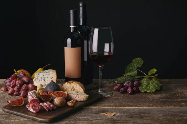 Garrafas e copo de vinho tinto, frutas e deliciosos queijos na mesa de madeira — Fotografia de Stock