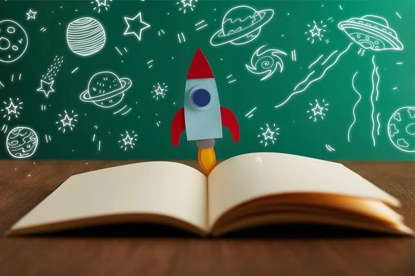 Open book with colorful rocket on wooden table with universe icons — Stock Photo