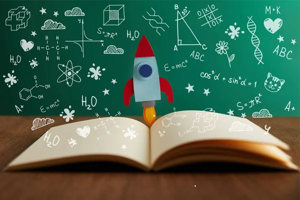 Відкрита книга з різнокольоровою ракетою на дерев'яному столі з математичними іконками — стокове фото