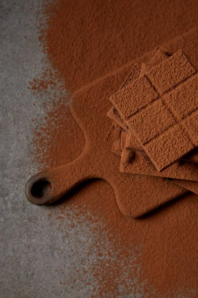 Вид сверху на сладкие вкусные кусочки шоколада с какао-порошком на доске для рубки на сером — стоковое фото