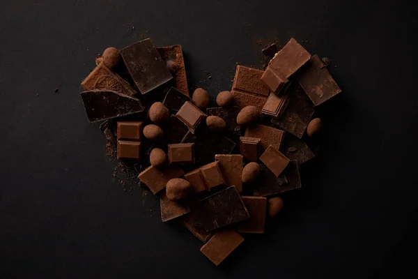 Vue de dessus des morceaux de chocolat gourmet sucré avec des noisettes disposées en forme de coeur sur noir — Photo de stock