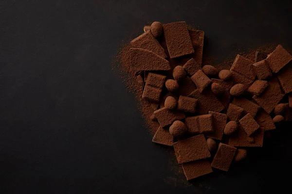 Vista dall'alto di deliziosi pezzi di cioccolato con noci e cacao in polvere disposti a forma di cuore su sfondo nero — Foto stock