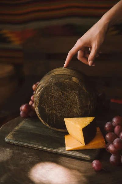 Tiro cortado de mulher segurando cabeça de queijo na tábua de corte com uvas — Fotografia de Stock