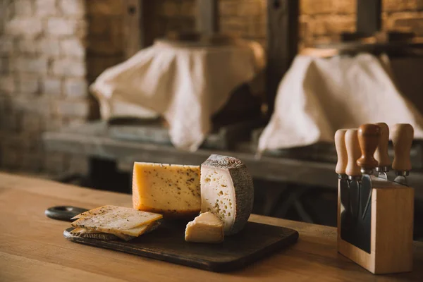 Primer plano de delicioso queso en rodajas en la tabla de cortar con varios cuchillos en el soporte - foto de stock