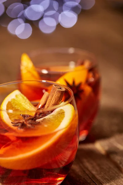 Vue rapprochée des boissons chaudes au vin chaud chaud avec des épices et des morceaux d'orange sur une table en bois avec des lumières bokeh floues sur fond — Photo de stock