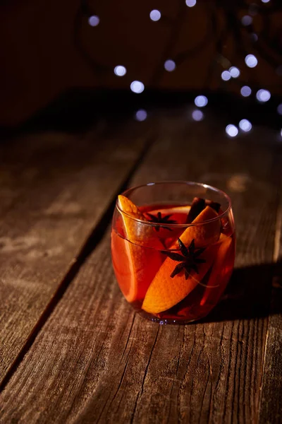 Close up view of hot mulled wine drink with orange pieces and anise stars on wooden surface with bokeh lights on background — Stock Photo