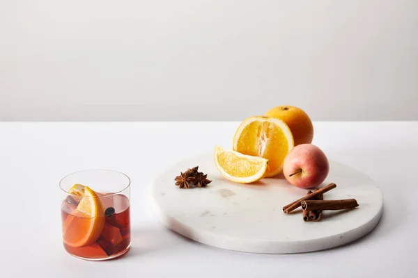 Nahaufnahme von Glühwein im Glas, Gewürzen, Orangen und Äpfeln auf weißer Oberfläche vor grauem Hintergrund — Stockfoto