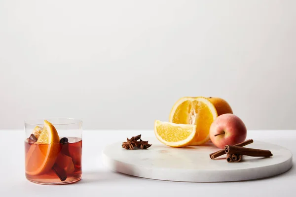 Vista de cerca del vino caliente caliente caliente caliente en vidrio, especias, naranjas y manzana en la superficie blanca sobre fondo gris - foto de stock
