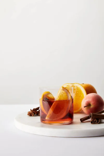 Близкий вид горячего глинтвейна в стакане, специи, апельсины и яблоко на белой поверхности на сером фоне — стоковое фото