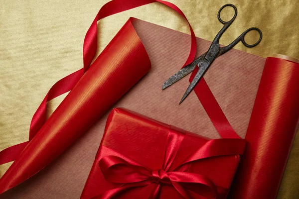 Vista superior de regalo envuelto, cintas y tijeras sobre fondo de papel de regalo rojo y dorado - foto de stock