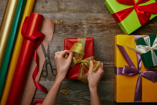 Cropped shot of woman tying ribbon on wrapped christmas present on wooden surface with other gifts — Stock Photo