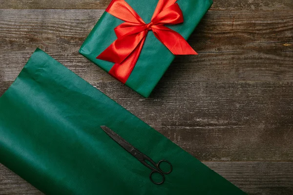 Top view of green wrapping paper, scissors and present with red ribbon on wooden background — Stock Photo