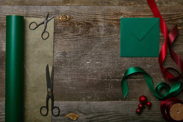 Tendido plano con sobre en blanco, tijeras, cintas verdes y rojas y papel de envolver en la superficie de madera, fondo de Navidad - foto de stock