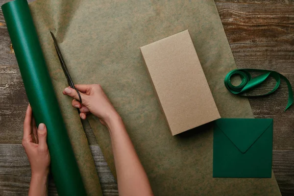 Teilansicht einer Frau, die Geschenkpapier mit Schere auf Holzoberfläche mit Geschenk und Umschlag schneidet — Stockfoto