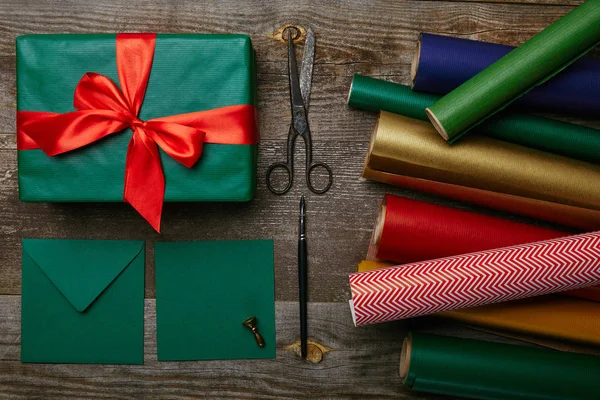 Cama plana con regalo de Navidad con cinta roja, papeles de embalaje, tijeras y sobres para la tarjeta de felicitación en la superficie de madera - foto de stock