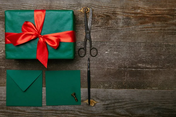 Posa piatta con regalo di Natale avvolto con nastro rosso, forbici e buste per biglietto di auguri su superficie di legno — Foto stock
