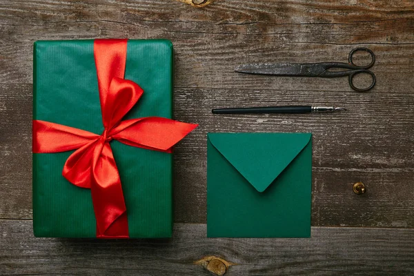 Posa piatta con regalo di Natale avvolto con nastro rosso, forbici e busta per biglietto di auguri su superficie di legno — Foto stock