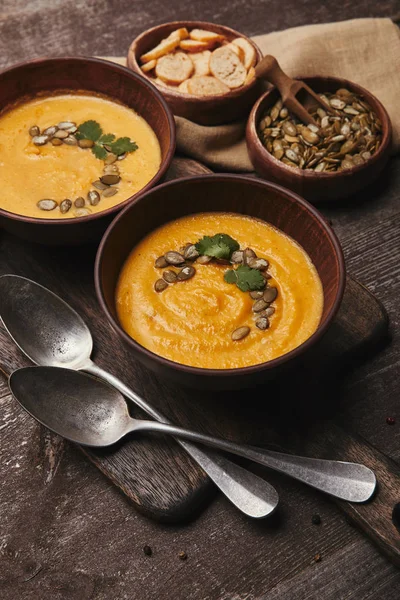 Vista de cerca de la deliciosa sopa de calabaza con semillas en cuencos y cucharas en mesa de madera - foto de stock