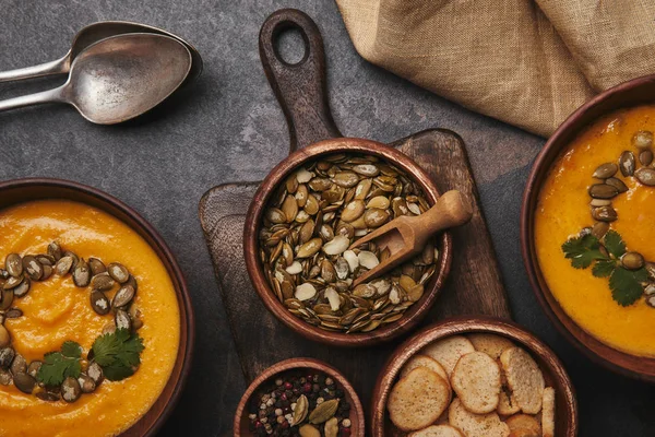 Vista superior de semillas de calabaza saludables, especias, rusks y sabrosa sopa de calabaza en cuencos - foto de stock