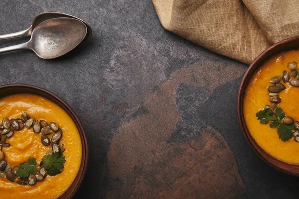 Вид зверху на органічний гарбузовий суп у мисках, ложках та мішковині на темній поверхні — стокове фото